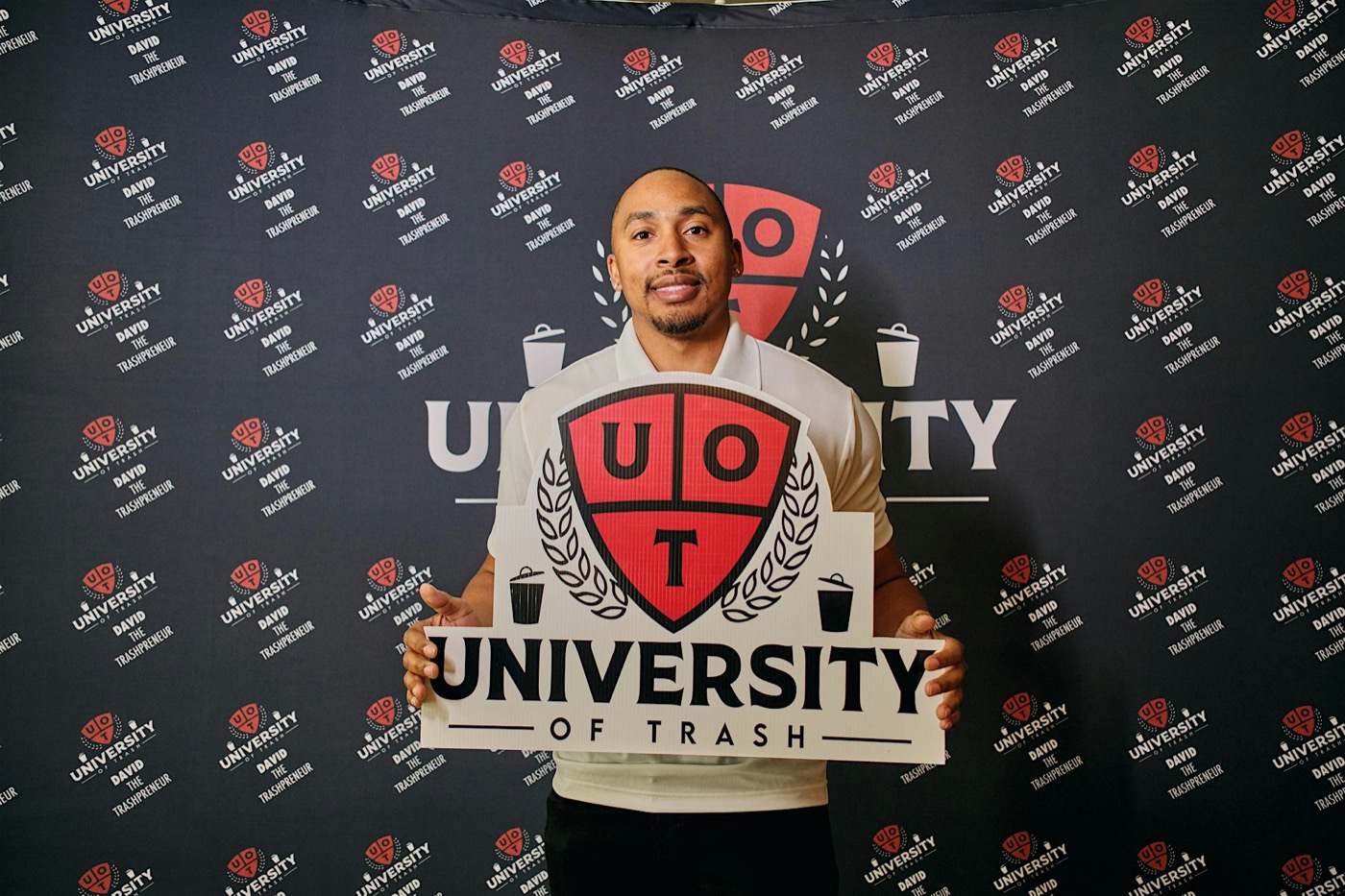 David Simes holding a large University of Trash crest sign in front of a branded step-and-repeat backdrop.